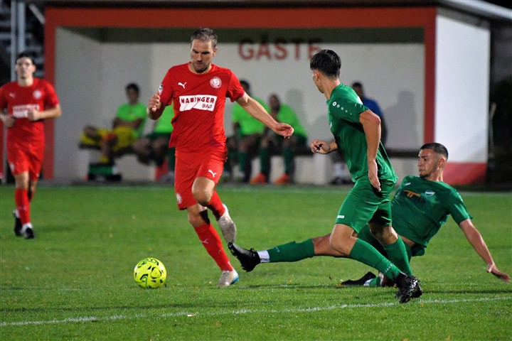 Fußballspieler in Rot dribbelt am Ball, Gegenspieler in Grün versucht zu grätschen